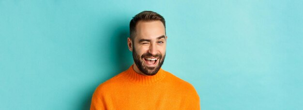 青い背景に対してオレンジ色のセーターで生意気な立っている笑顔のカメラでウィンクしている自信を持ってハンサムな男