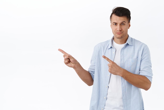 Handsome confident masculine man in shrit pointing fingers left and look persuasive and assured camera make final choice recommend service product or shopping site white background