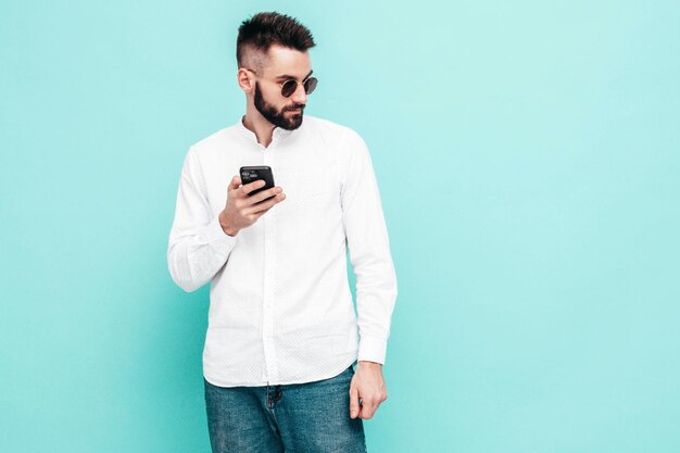 잘생긴 자신감 모델 셔츠와 청바지를 입은 섹시하고 세련된 남자 스튜디오의 파란색 벽 근처에서 포즈를 취하는 패션 힙스터 남성 스마트폰 들고 휴대폰 화면 보기 격리된 앱 사용