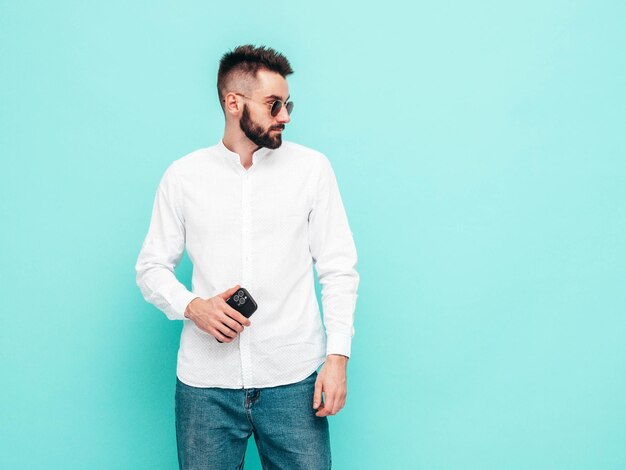 Handsome confident modelSexy stylish man dressed in shirt and jeans Fashion hipster male posing near blue wall in studio Holding smartphone Looking at cellphone screen Using apps Isolated