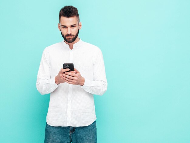 Handsome confident modelSexy stylish man dressed in shirt and jeans Fashion hipster male posing near blue wall in studio Holding smartphone Looking at cellphone screen Using apps
