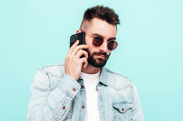 Free photo handsome confident modelsexy stylish man talking at smartphone fashion hipster male posing near blue wall in studio holding phone with cellphone in sunglasses isolated