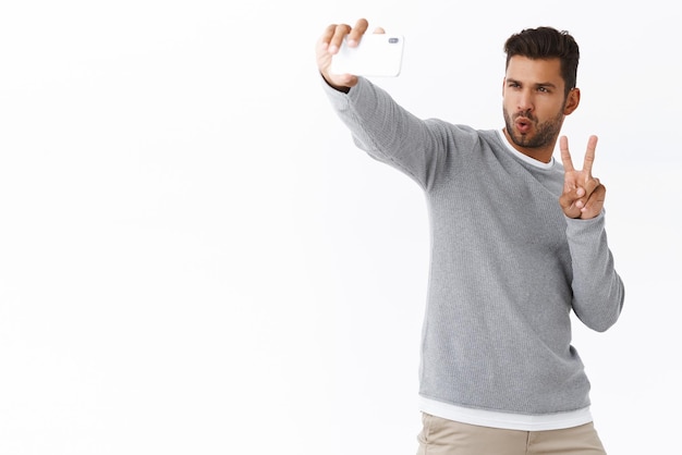 Free photo handsome confident sexy guy with bristle in grey sweater taking selfie mobile camera posing with folded lips during concert or party make peace gesture hold smartphone with raised arm