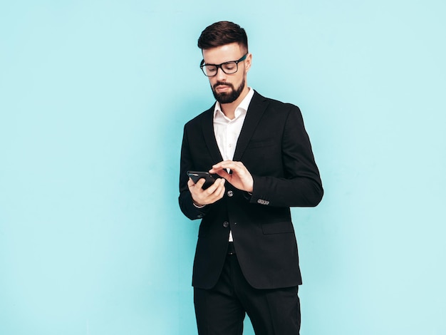 Handsome confident stylish hipster lambersexual model Sexy modern man dressed in elegant black suit Fashion male posing in studio Holding smartphone Looking at cellphone screen Using apps