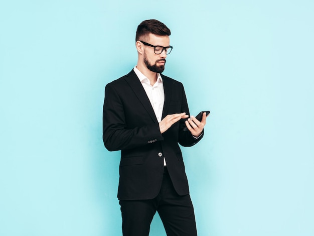 Handsome confident stylish hipster lambersexual model Sexy modern man dressed in elegant black suit Fashion male posing in studio Holding smartphone Looking at cellphone screen Using apps