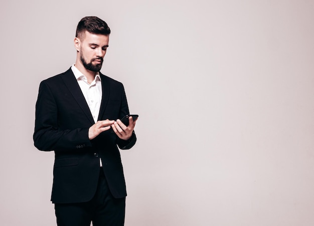 잘 생긴 자신감 있는 세련된 힙스터 램버섹슈얼 모델 우아한 검은 양복을 입은 섹시한 현대 남성 스튜디오에서 포즈를 취하는 패션 남성 스마트폰 들고 휴대폰 화면 보기 앱 사용