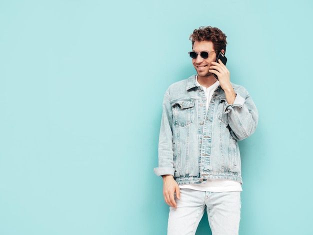 Handsome confident stylish hipster lambersexual model with curly hairstyle Sexy man dressed in jeans jacket Fashion male holding phone With cellphone Talking at smartphone