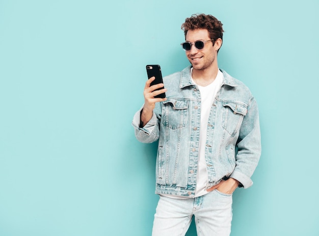 Handsome confident stylish hipster lambersexual model with curly hairstyle Sexy man dressed in jeans jacket Fashion male holding smartphone Looking at cellphone screen Using apps