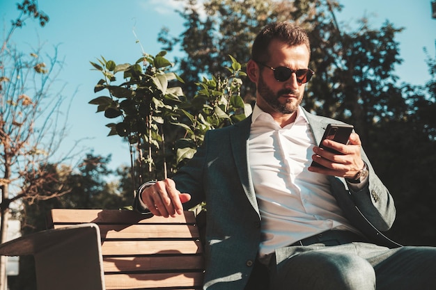 Modello lambersexual bello fiducioso elegante hipster uomo moderno vestito con un abito elegante moda maschile in posa per strada nella città europea in occhiali da sole seduto in panchina guardando smartphone