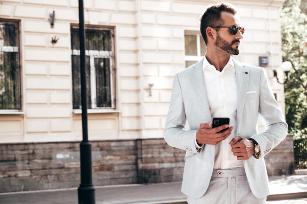 잘 생긴 자신감이 세련된 힙스터 램버섹슈얼 모델흰색 양복을 입은 현대 남성 일몰 유럽 도시의 거리에서 포즈를 취하는 섹시한 패션 남성 선글라스를 끼고 스마트폰을 보고