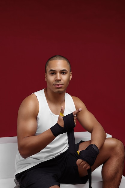 Handsome confident young African businessman with muscular shoulders wearing black shorts and white tank top tying boxing bandages for workout using punching bag, training after work day at office