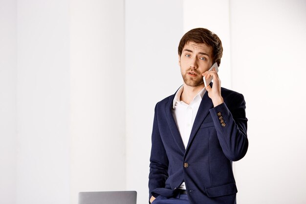 Красивый уверенный молодой бизнесмен, стоящий возле офисного стола и разговаривающий по телефону