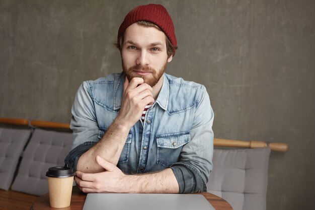 Handsome confident young Caucasian bearded freelancer in jeans jacket and hat