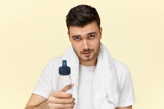 Handsome confident young European male with bristle posing with white towel around his neck, drinking water from plastic cup after physical training