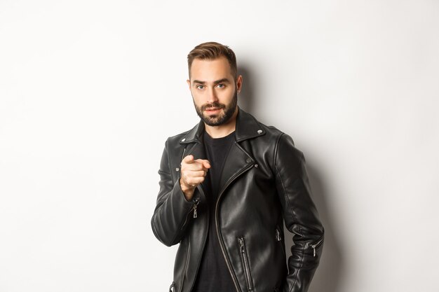 Handsome and cool bearded guy pointing finger at camera, wearing leather jacket, standing sassy