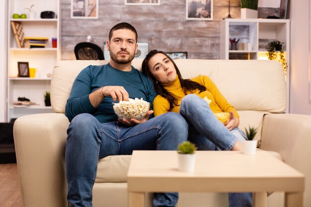 ポップコーンを食べて、リビングルームのソファでテレビを見ている自宅でハンサムなカップル