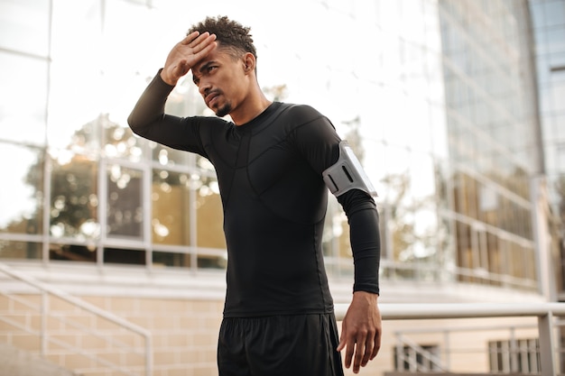 Handsome curly dark-skinned man in black long-sleeved t-shirt and shorts touches his face