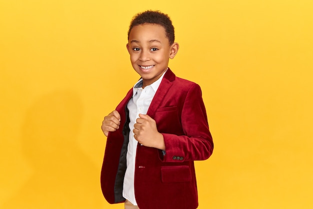 Handsome cute African American little fashionmonger posing isolated having confident facial expression, smiling, adjusting trendy crimson velvet jacket.