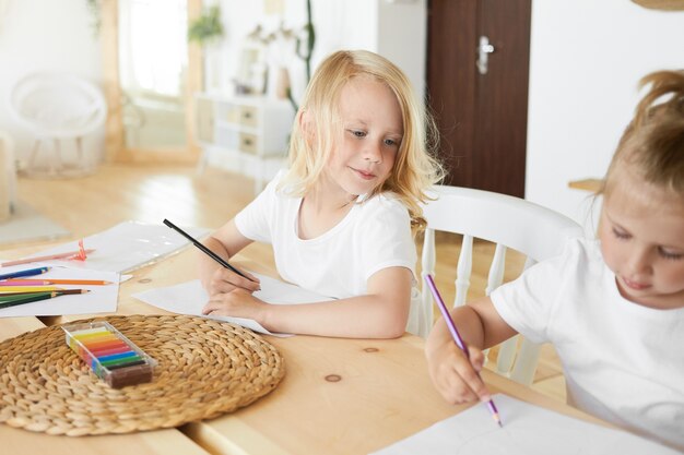 Handsome cute schoolboy with loose blond hair holding pencil having curious facial expression, looking at his baby sister who is sitting next to him, drawing something on blank white sheet of paper