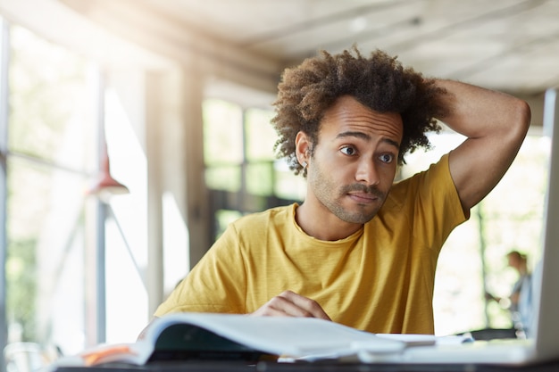 Handsome dark-skinned curly stylish male dressed in T-shirt scratching his head while looking at laptop having some problems with studying