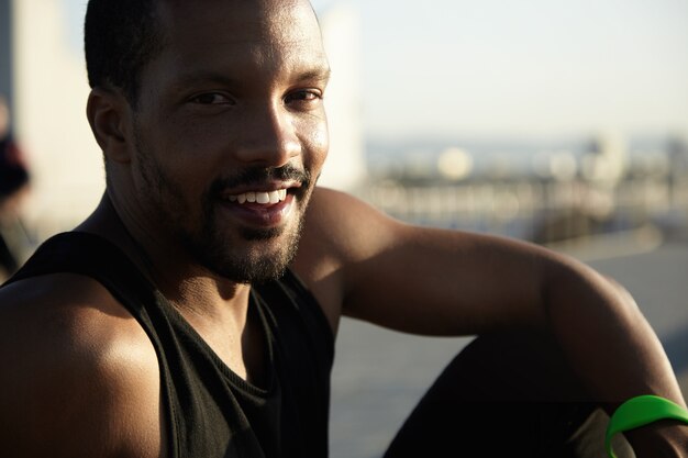 Handsome dark-skinned runner resting at seaside after activity in open air sitting on embankment, enjoying sunset on summer evening. Cheerful student spending his vacations exercising outdoors