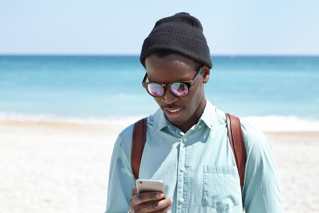Handsome dark-skinned student in fashionable wear spending free time after college by the sea, having nice walk along beach, messaging friends online. People, lifestyle and modern technology