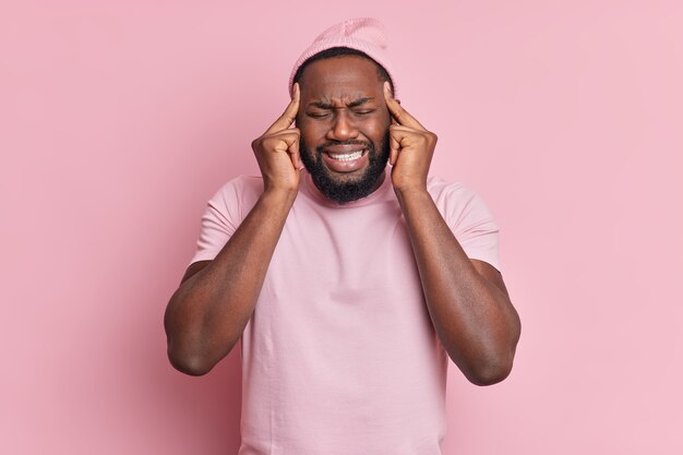 Handsome displeased man with thick beard suffers from unbearable migraine keeps fingers on temples to reveal pain clenches teeth wears casual t shirt and hat poses over pale pink wall