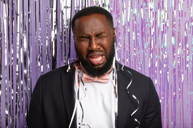 Handsome dissatisfied black Afro American man with desperate look, cries from depression, wears elegant suit, poses against purple wall with tinsel. Negative human emotions, spoiled party