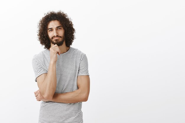 handsome Eastern male coworker in trendy striped t-shirt, leaning head on fist while standing with half-crossed fingers