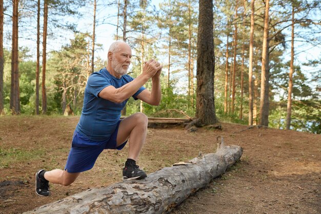 野生の自然の中で有酸素運動をしているスポーツ服を着ているひげを持つハンサムなエネルギッシュな年配の男性。走る前に足を丸太につけ、脚の筋肉を鍛える、うれしそうな自信に満ちた表情の老人