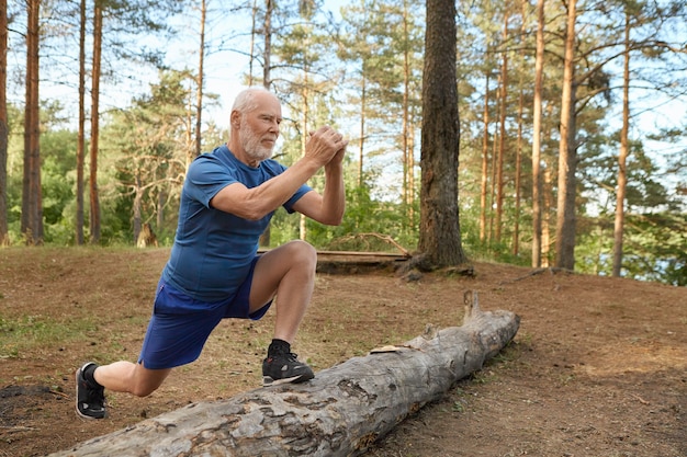 Handsome energetic senior man with beard wearing sports clothes doing cardio routine in wild nature. Elderly man having joyful confident look keeping foot on log, training leg muscles before run