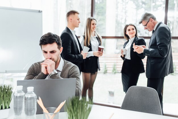 Handsome enterpreneur with laptop sitting at modern office and thinking, while his colleagues have a break time with coffe.