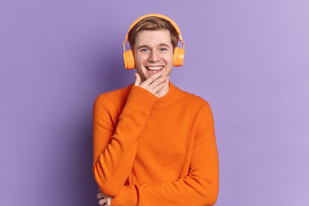 Handsome European guy smiles gladfully expresses positive emotions listens audio track via stereo headphones wears orange jumper 