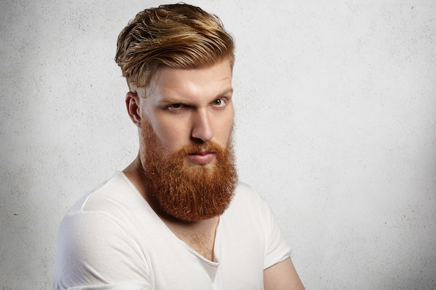   handsome fashionable man with long red beard and trendy hairstyle having serious and displeased facial expression scowling and frowning while standing against concrete wall  