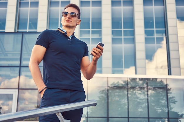 Красивый модный мужчина со стильной стрижкой в солнцезащитных очках, одетый в черную футболку и брюки, держит смартфон, стоит в современном городе на фоне небоскреба.