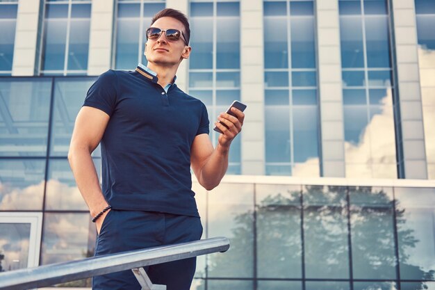 A handsome fashionable man with a stylish haircut in sunglasses, dressed in a black t-shirt and pants, holds the smartphone, stans in the modern city against a skyscraper.