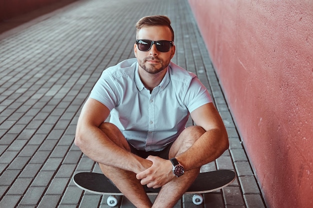 Free photo handsome fashionable skater guy in sunglasses dressed in a white shirt and shorts sitting on a skateboard under the bridge, looking at a camera.