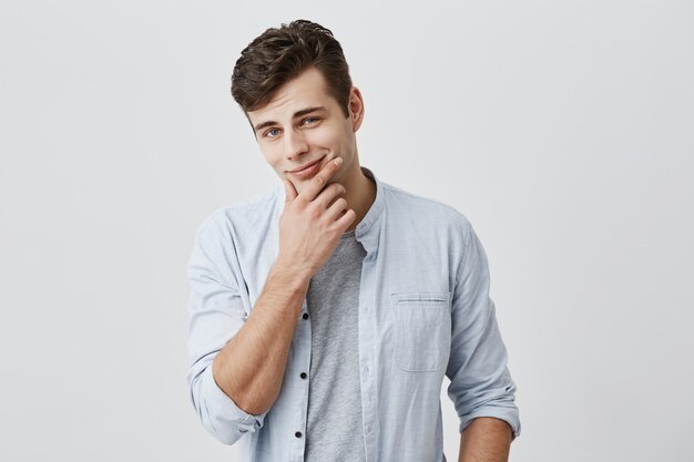 Handsome flirting attractive caucasian male in light blue shirt with dark hair and blue eyes, looking  with appeal and pleasant smile, touching chin,making tonight plans. Face expression