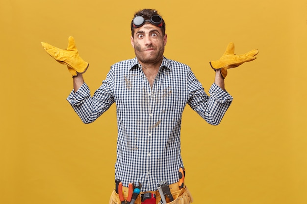 Handsome foreman wearing checkered shirt, goggles and protective gloves having belt with tools shrugging his shoulders not knowing where breakage is. Manual worker having doubts while working