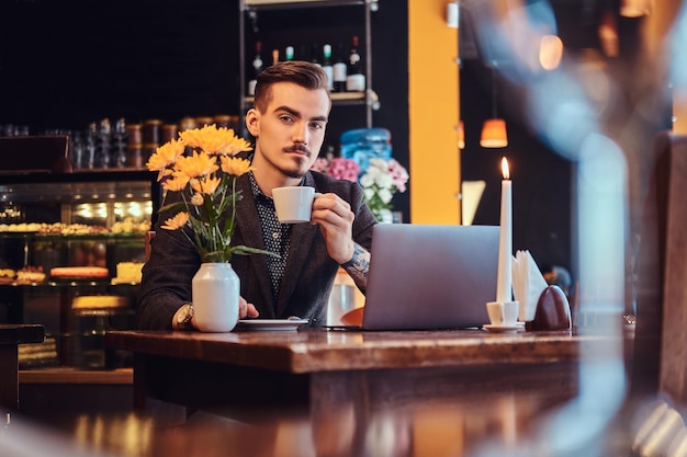 Handsome freelancer man with stylish beard and hair dressed in a black suit sitting at a cafe with an open laptop and holds a cup of coffee, looking at a camera.