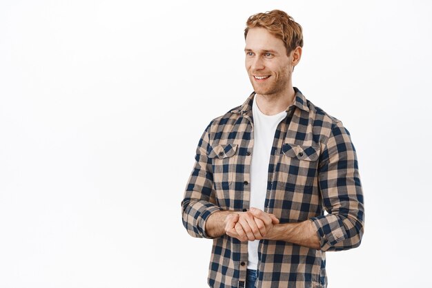 Handsome friendly-looking redhead man, holding hands together in polite poses, smiling and looking aside at client or customer, offer his assistance, helping you, standing over white wall