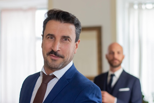 Handsome gay in official suit looking at camera. Front view of bearded Caucasian man smiling at camera, getting dressed for wedding ceremony with lover. LGBT, love, marriage concept