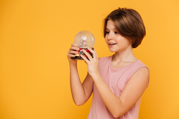 Handsome girl holding and looking at snow globe isolated over yellow