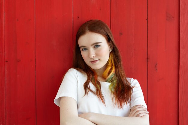 Handsome girl with colored strands in ginger long hair wearing white top looking with folded arms. Half body shot of green-eyed student female with crossed arms