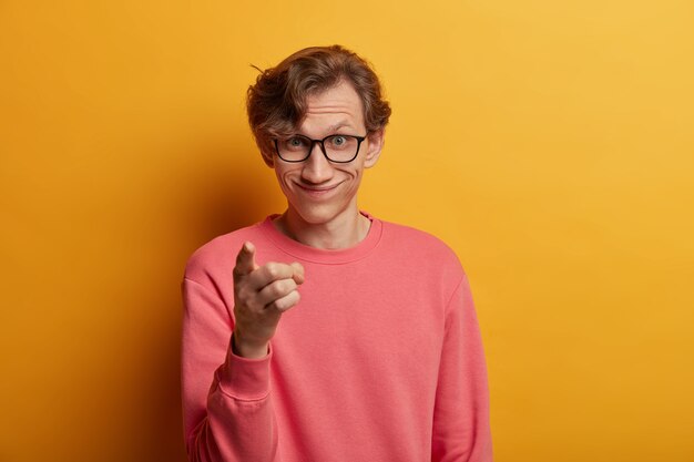 Handsome glad man giggles positively, selects someone, points fore finger directly , expresses choice, chooses something in shop to buy, wears rosy jumper, isolated on yellow  wall