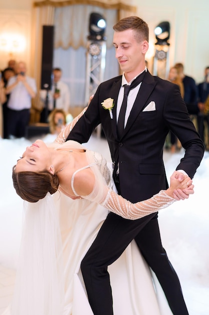 Handsome groom in black stylish suit holding hands of bride woman in beautiful wedding dress while dancing in the restaurant Attractive bride girl in long sleeves dress Loving married couple