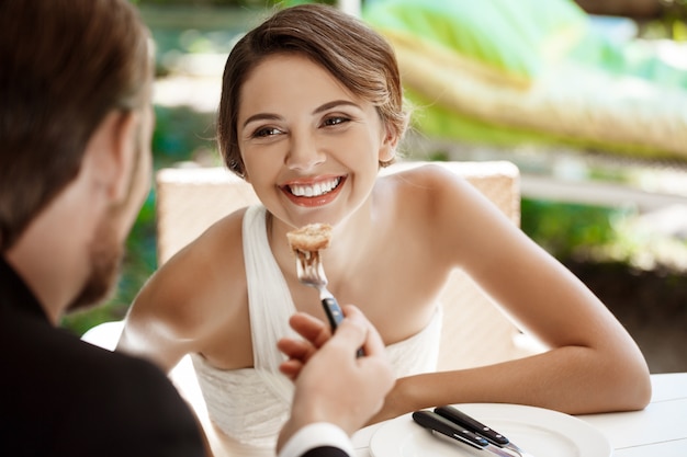 Handsome groom feeding his bride with croissant in cafe.