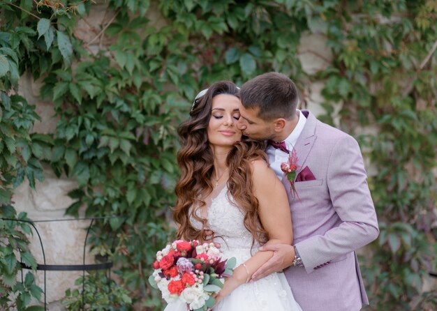 Handsome groom is kissing beautiful bride outdoors dressed in fashionable wedding attire
