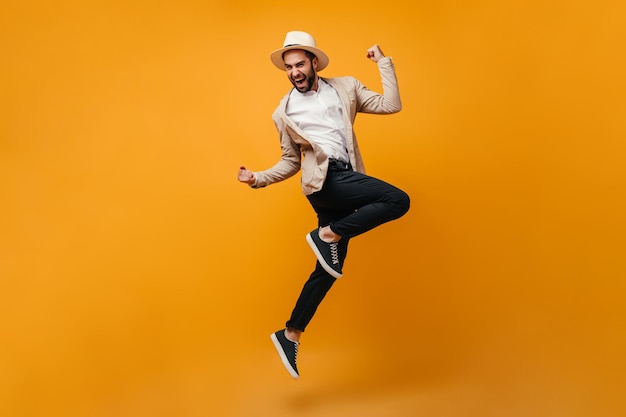 Free photo handsome guy in black pants and beige jacket jumps on orange background