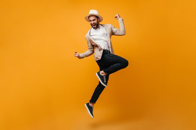 Handsome guy in black pants and beige jacket jumps on orange background